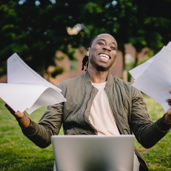 happy african american man holding sheets of paper in front of laptop